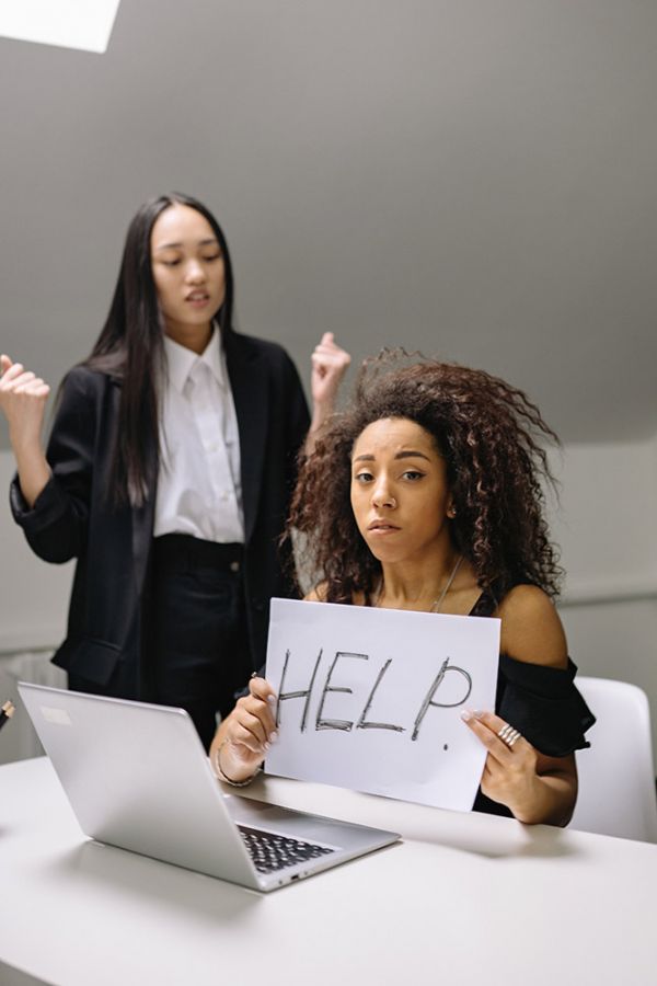 worried looking young woman holding a help sign being bullied by another young woman standing behind her