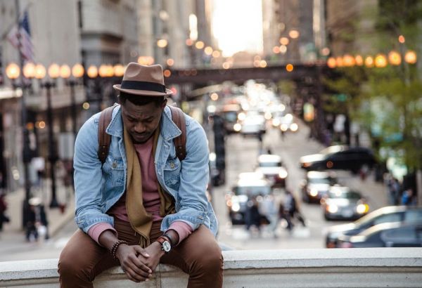 photo of a man in a leather jacket looking down while sitting on a ledge in a city